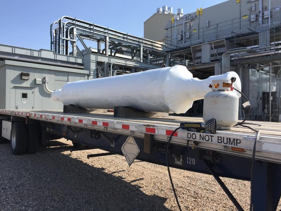 White industrial tank on a flatbed trailer at a plant, with refinery piping in the background.