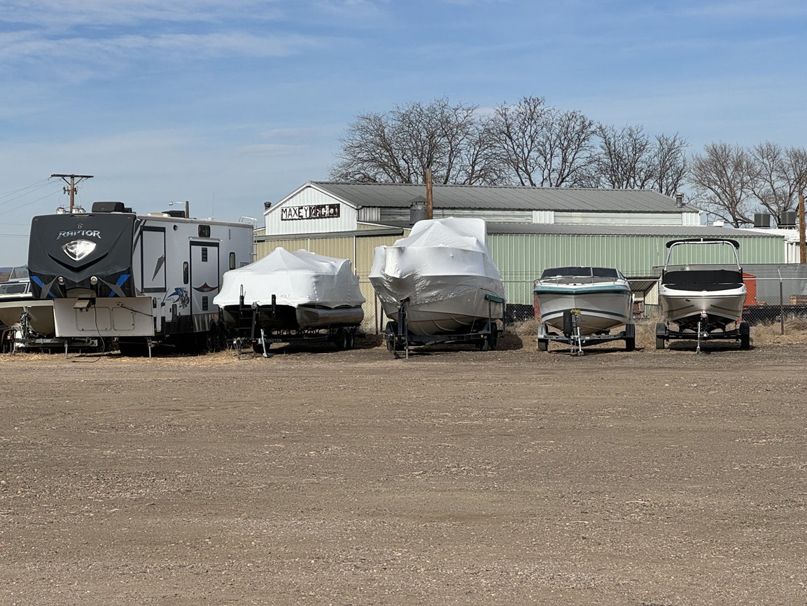 Several small boats stored in a gravel lot beside a shed under a blue sky