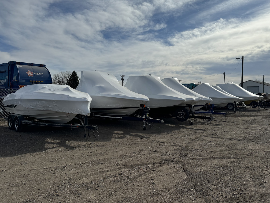 Covered boats on trailers parked in a gravel lot under a cloudy sky