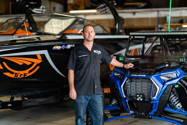Man standing beside a blue off-road vehicle and black race boat in a garage workshop
