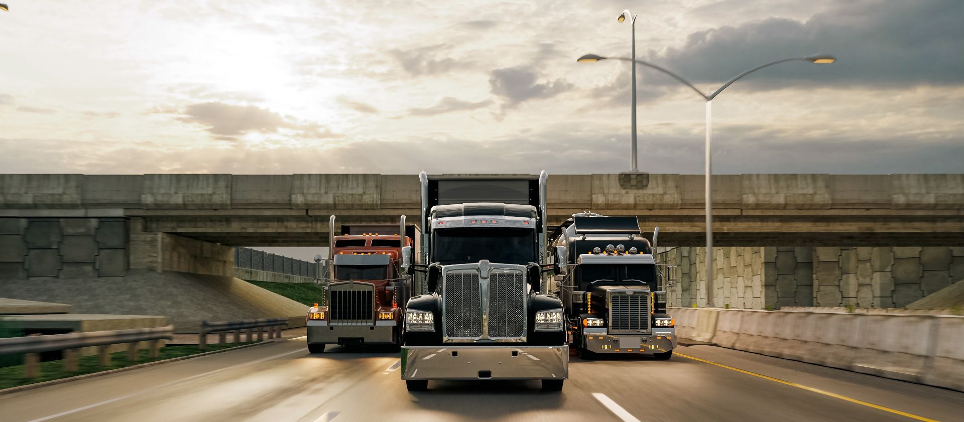 Three semi trucks driving on a highway under an overpass at sunset