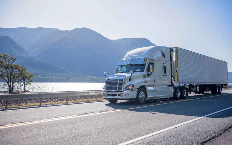 White semi-truck driving on a highway beside a lake and mountains on a sunny day