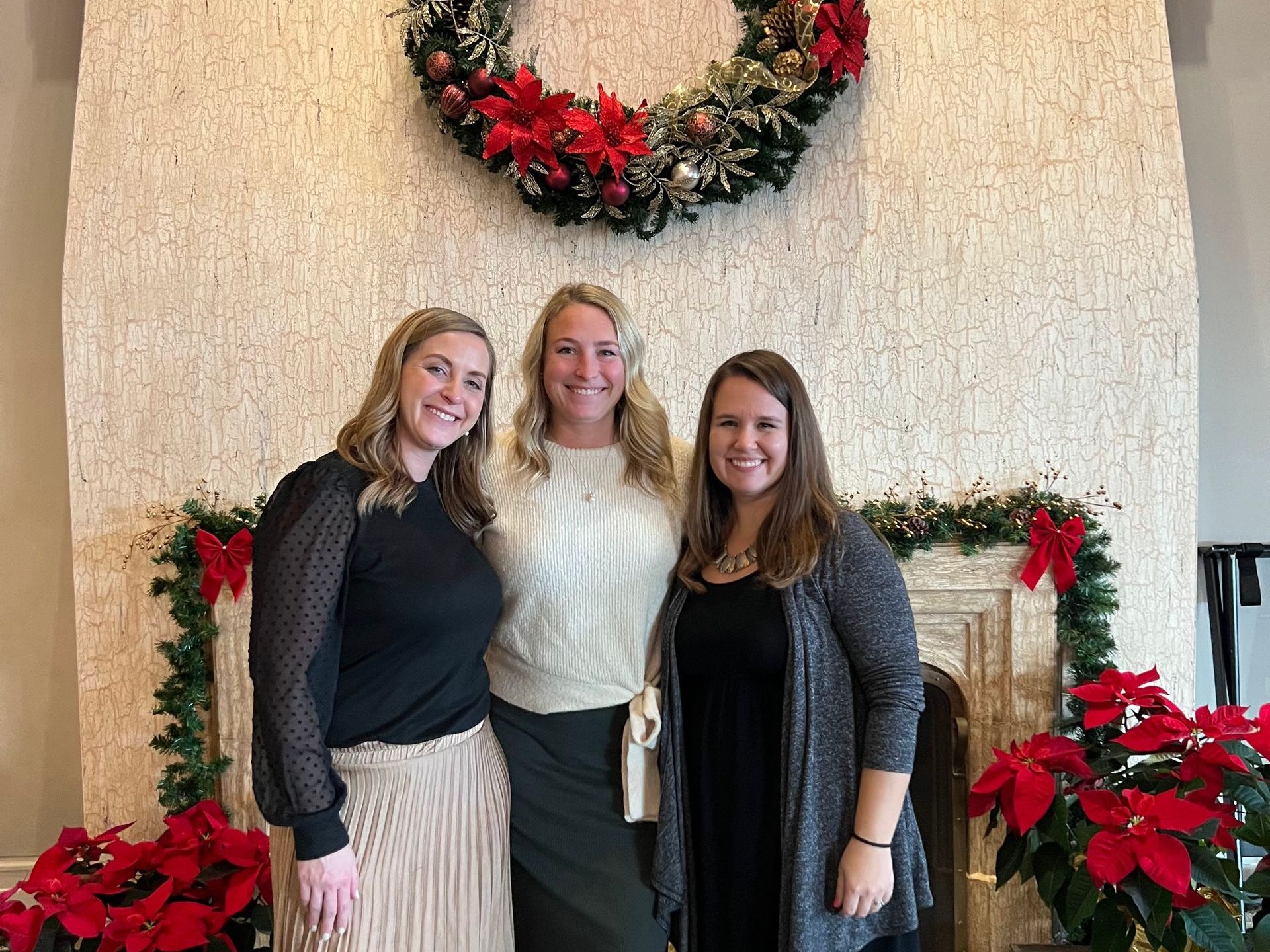 Three women pose in front of a fireplace decorated with a wreath, garland, and poinsettias.