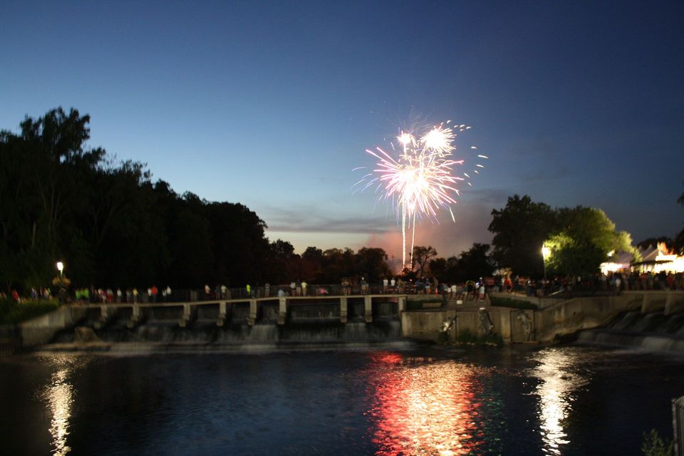 Fireworks exploding over a river at dusk; people watch from a dam.