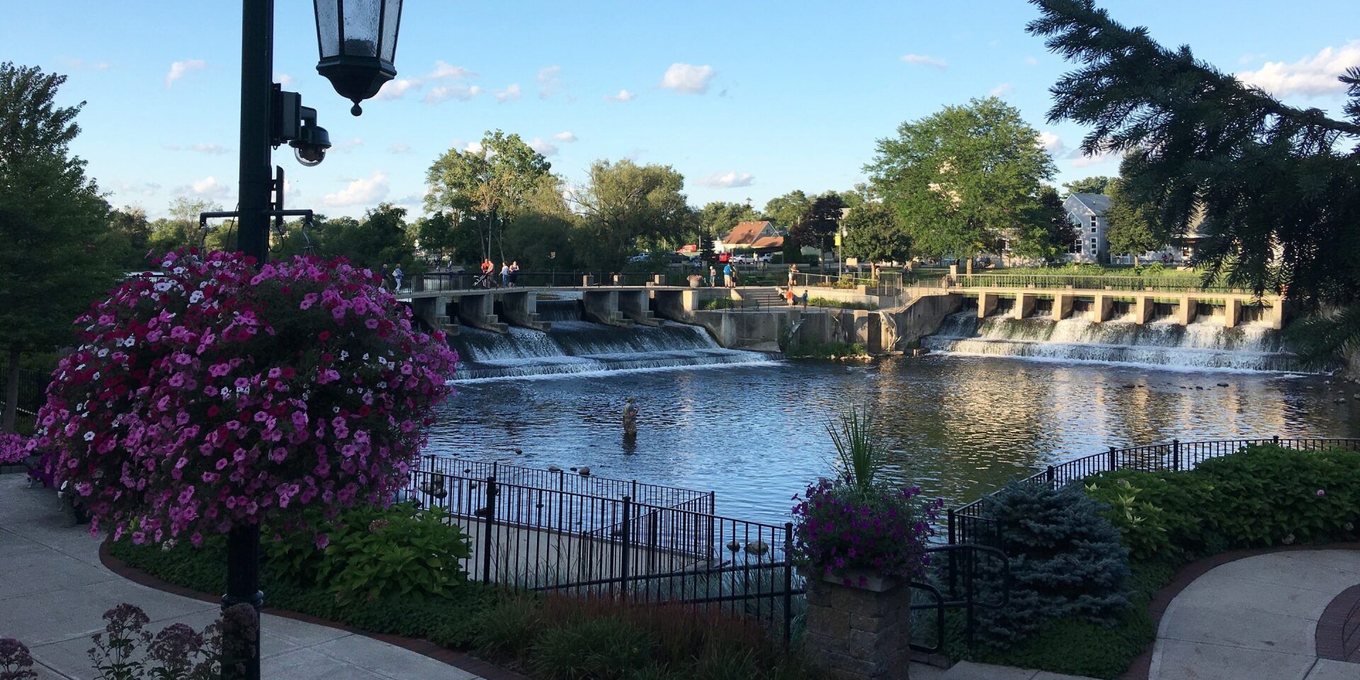 Scenic view of a river with a waterfall, surrounded by trees and a park with flowers.