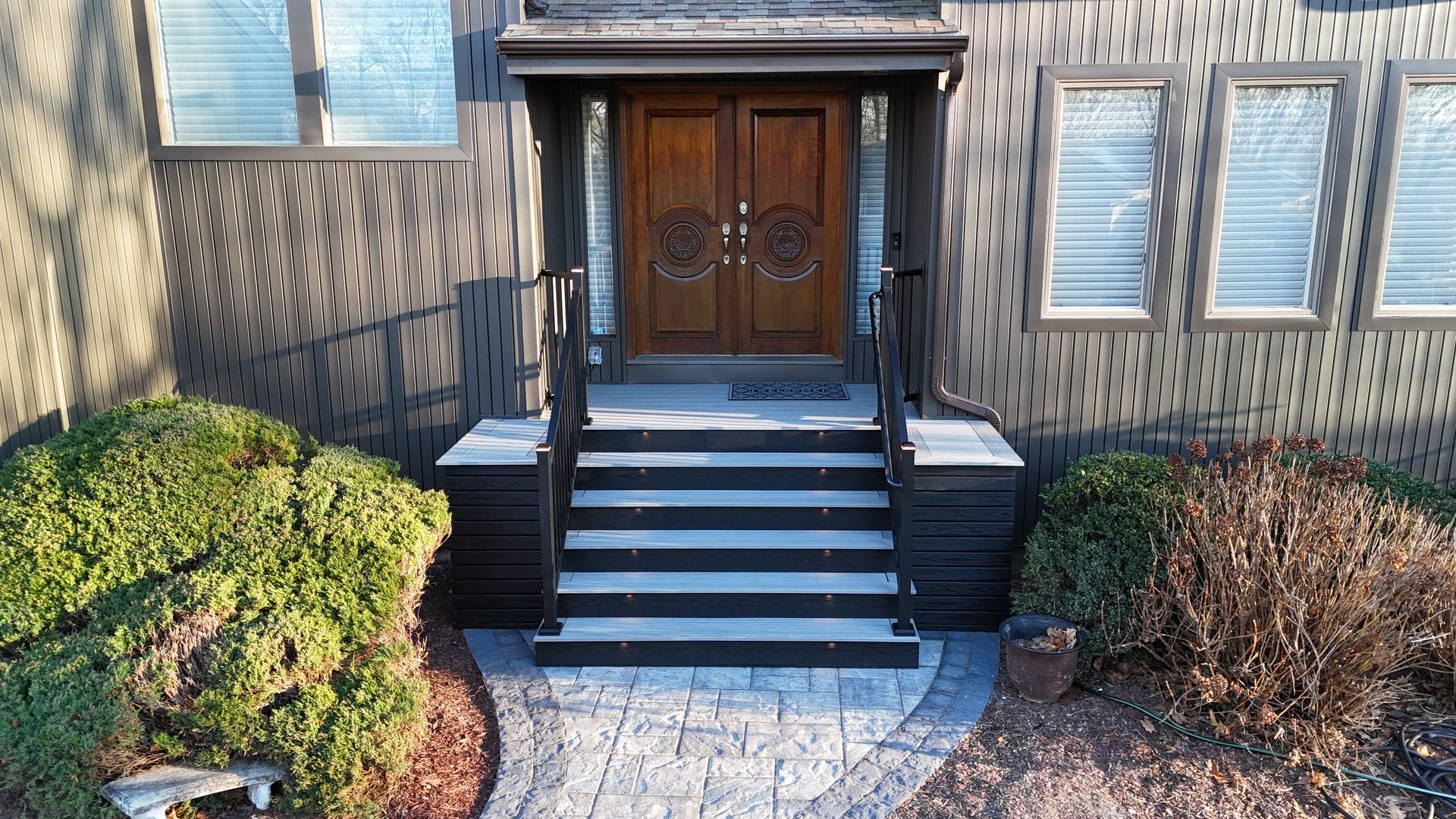 Front entrance with steps and double wooden doors. Dark siding, stone path, and greenery.