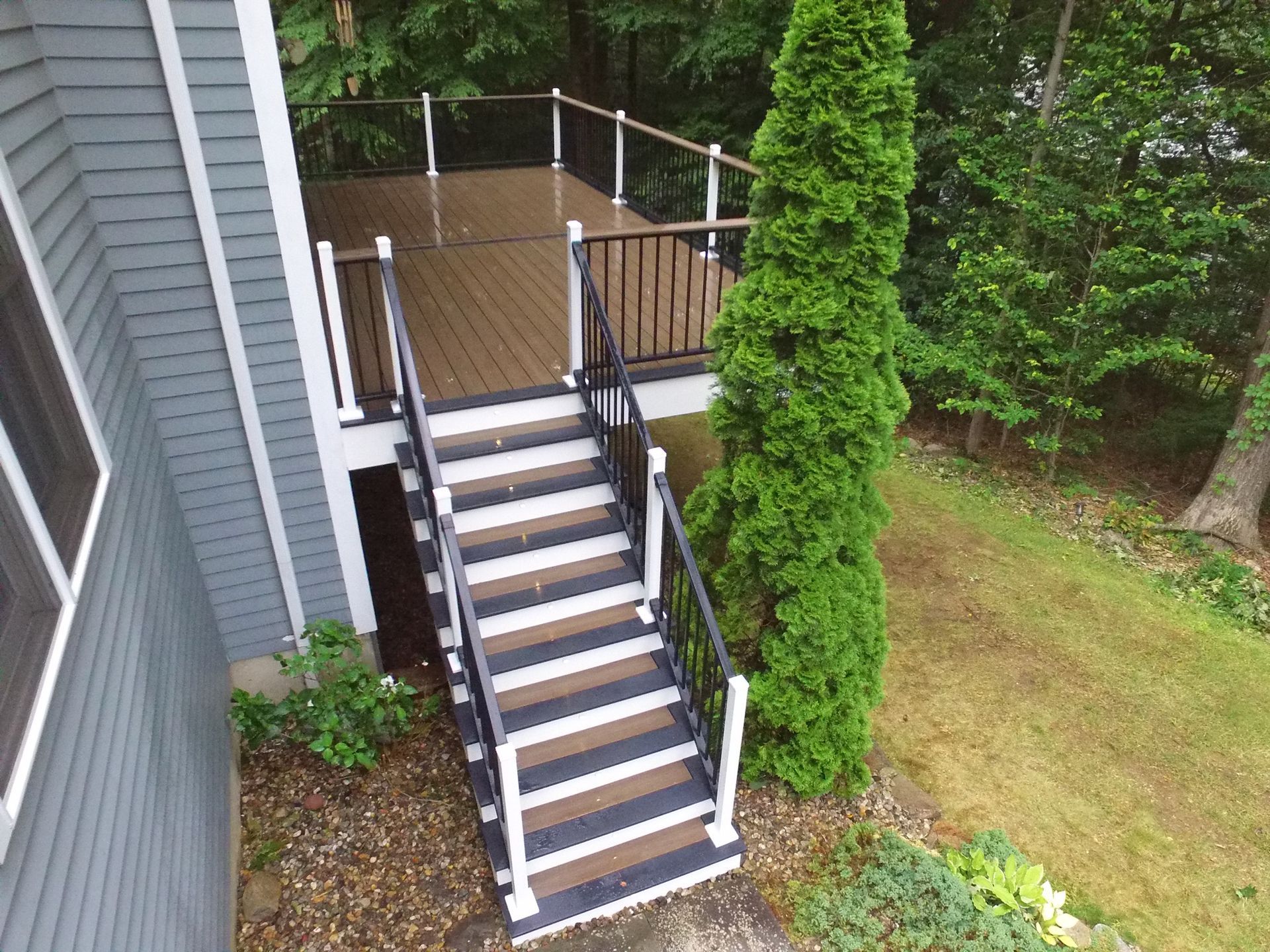 Deck with stairs, brown and white steps and railing, gray siding, surrounded by greenery.