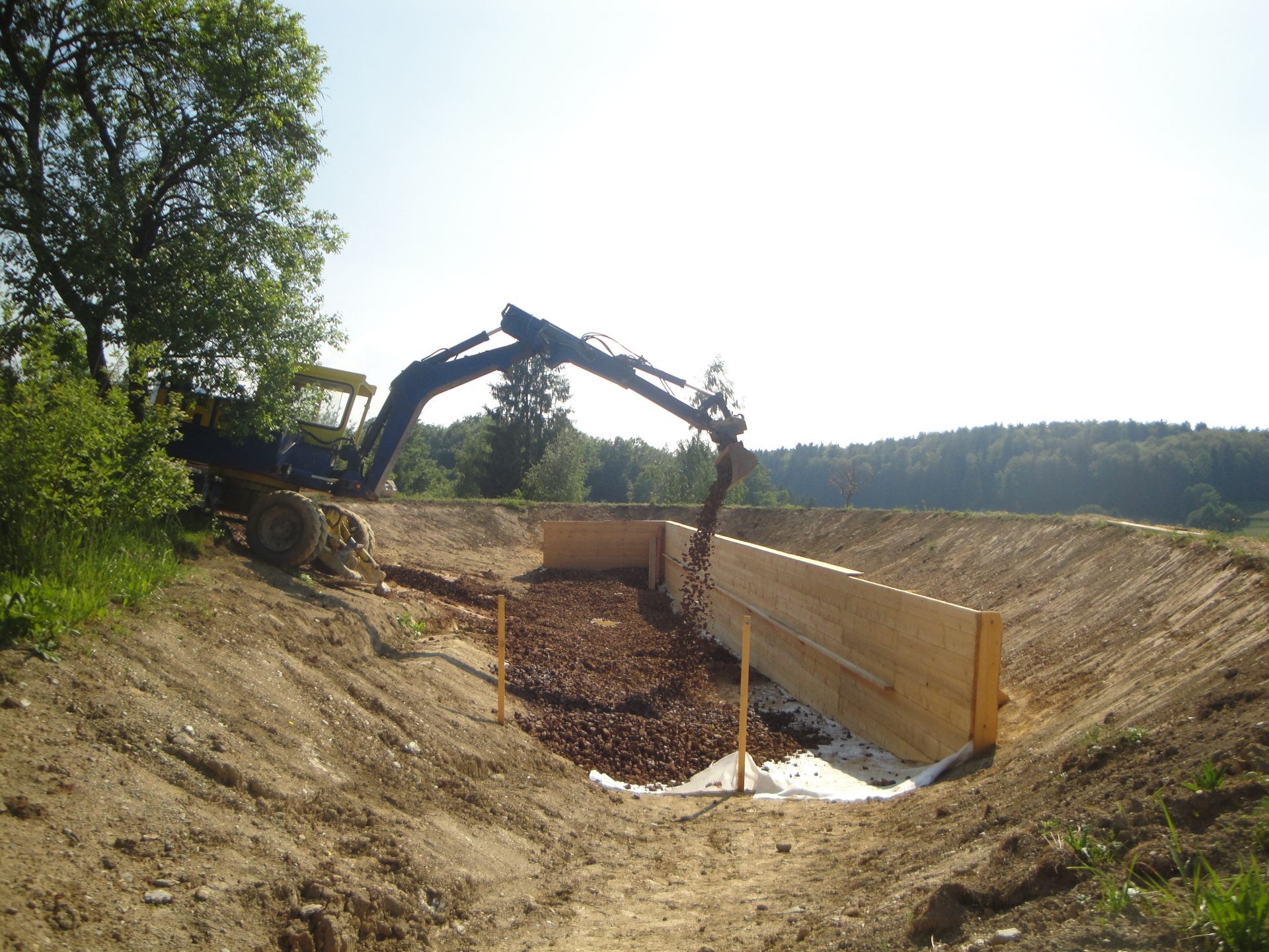An excavator works in a dirt pit, placing long wooden planks against an earthen wall lined with white material.