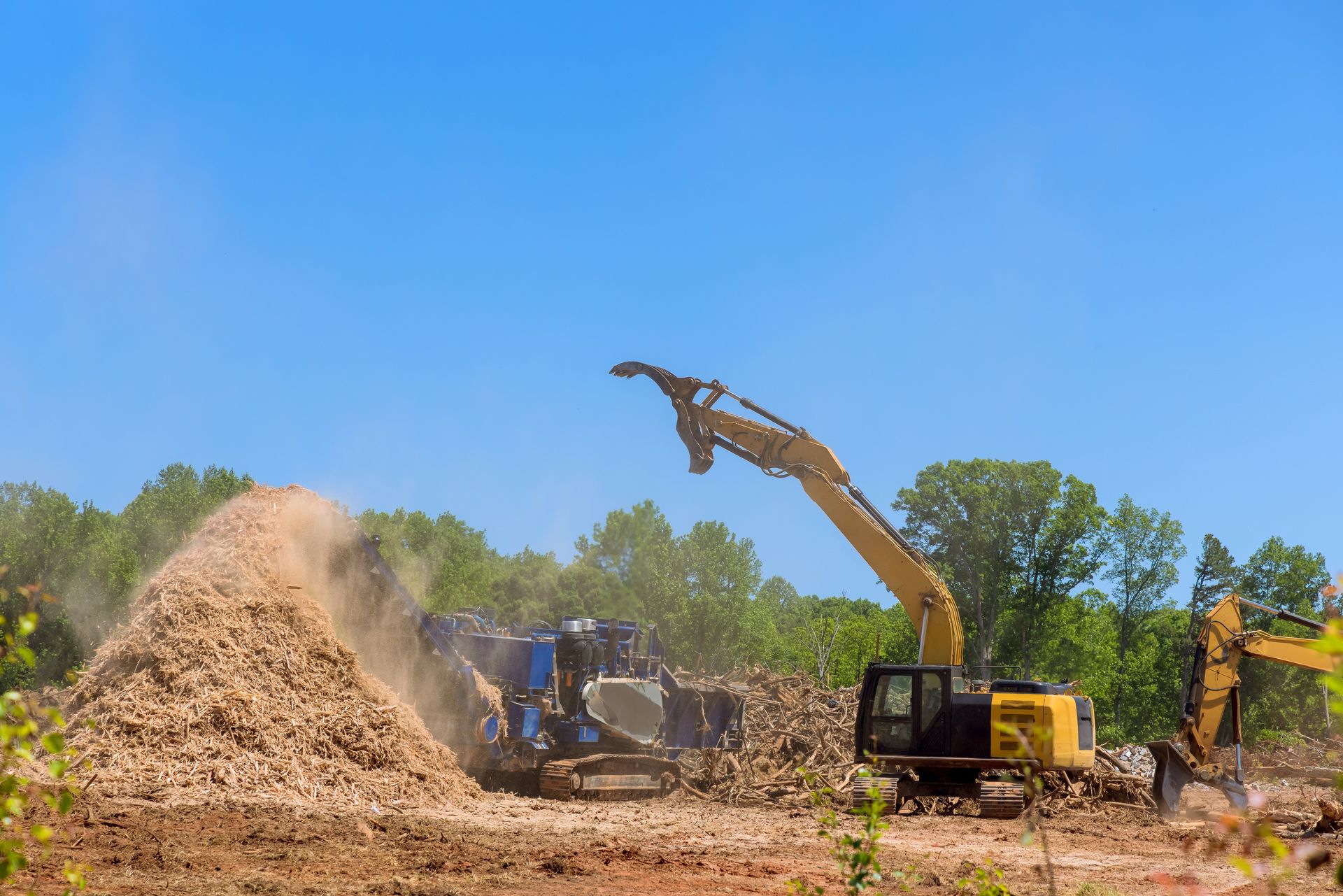Heavy machinery works at a construction site, processing debris into a large pile under a clear blue sky.