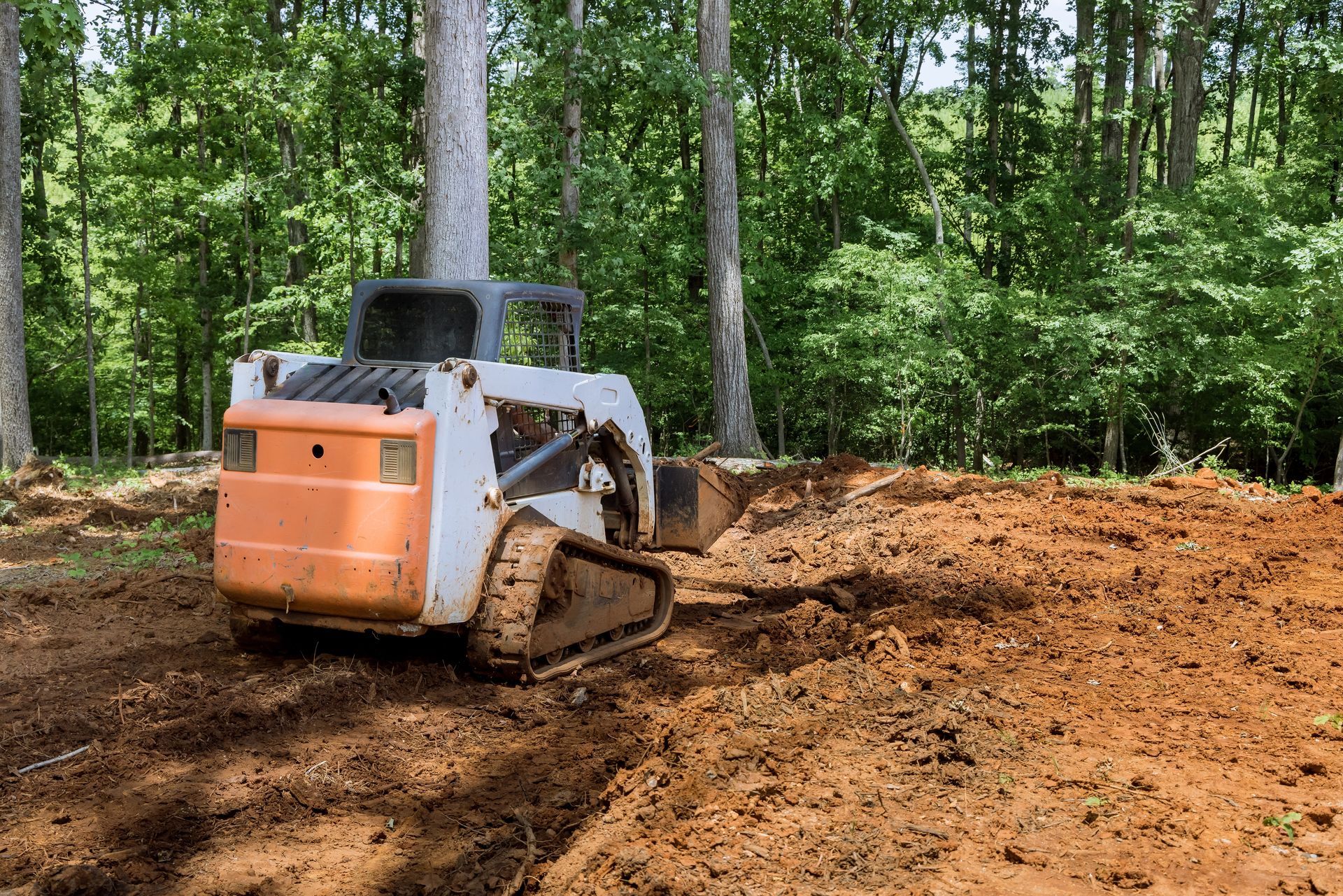 A white and orange tracked skid-steer loader moving dirt on a wooded construction site.