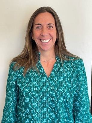 Smiling person in a teal patterned blouse standing against a plain light wall