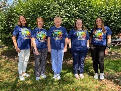 Five people standing outdoors on grass, wearing matching blue shirts in front of green trees.