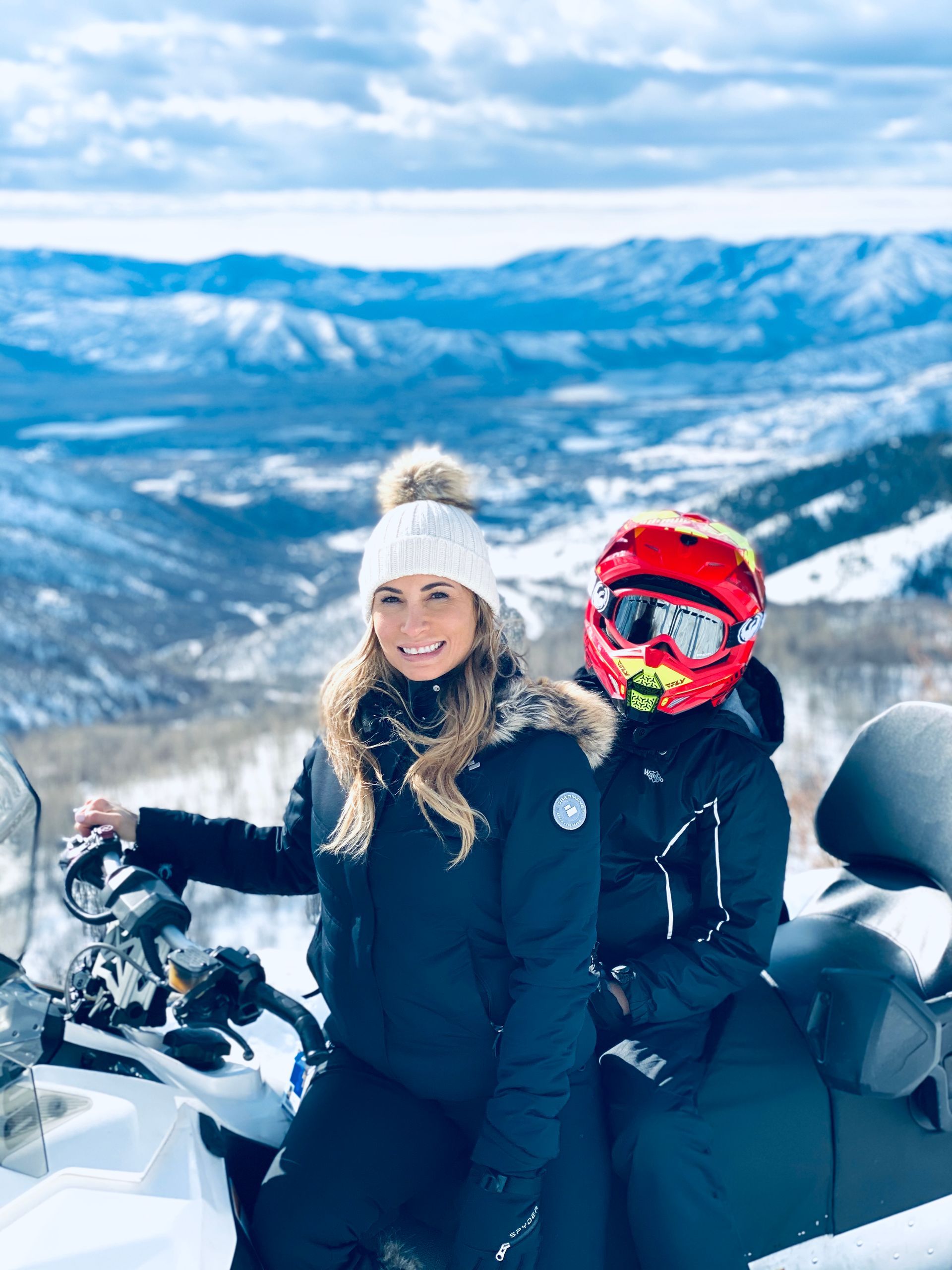 Two people in winter gear on a snowmobile, posing on a snowy mountain overlook.