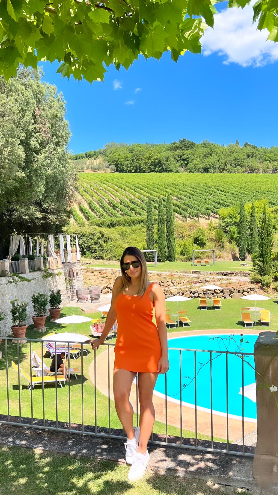 Woman in orange dress standing by a pool in a sunny garden with vineyard hills behind her