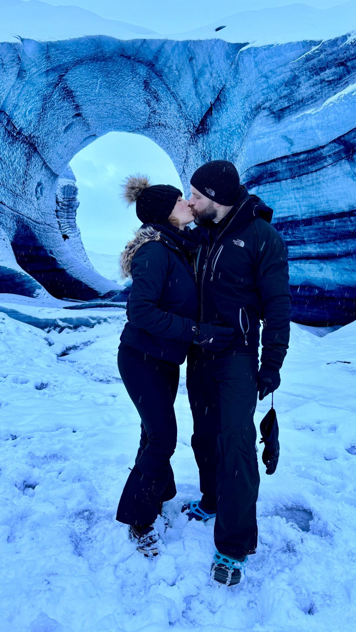 Two people kissing in snowy terrain in front of a large icy wall and circular opening