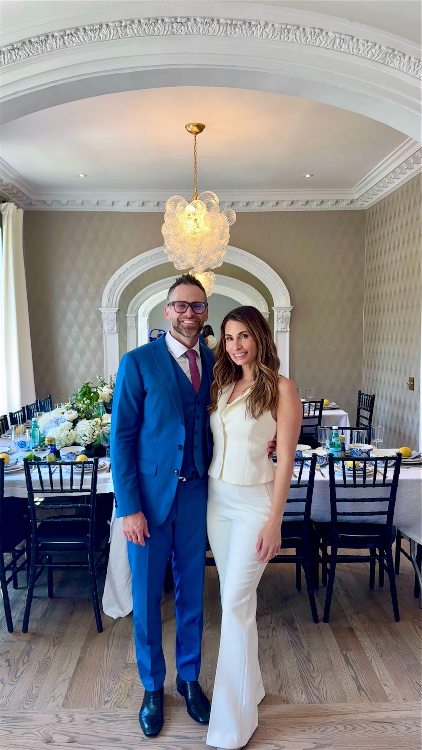 Couple posing in formal attire under a chandelier in an elegant dining room