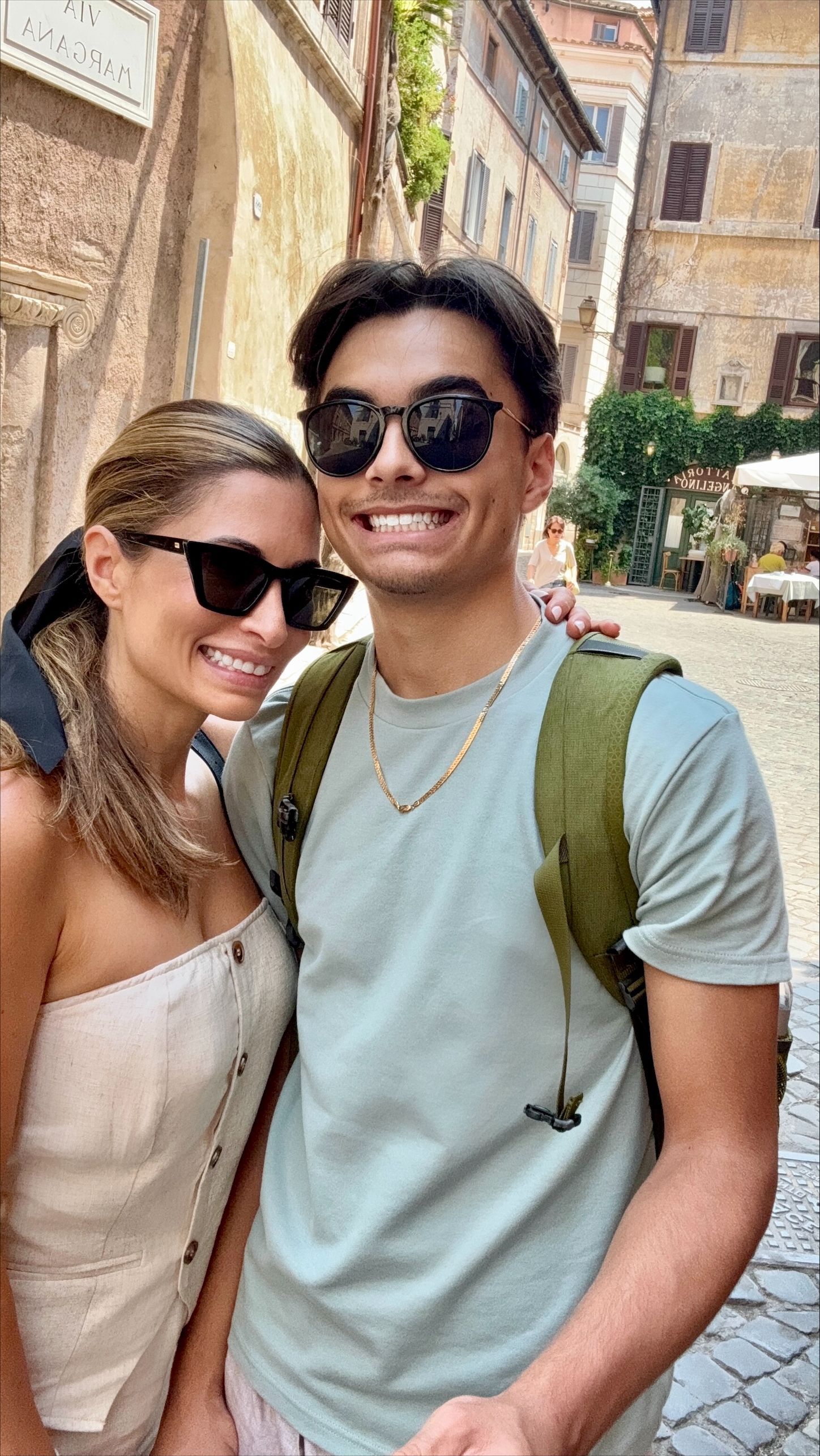 Two smiling people wearing sunglasses pose in a sunlit stone street, with one carrying a green backpack.