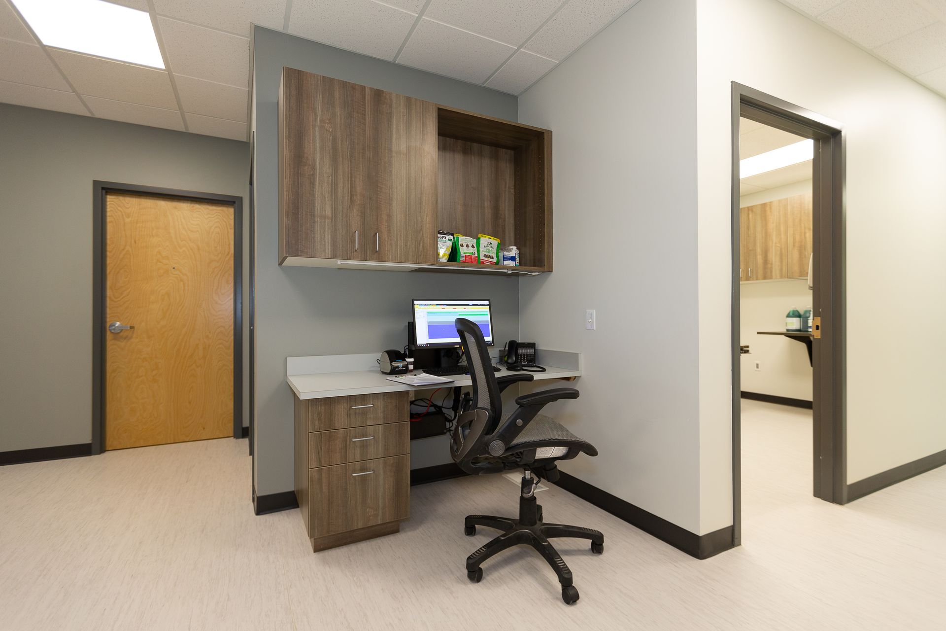 Empty office cubicle with wood cabinets, a desk, computer, and black swivel chair in a hallway.