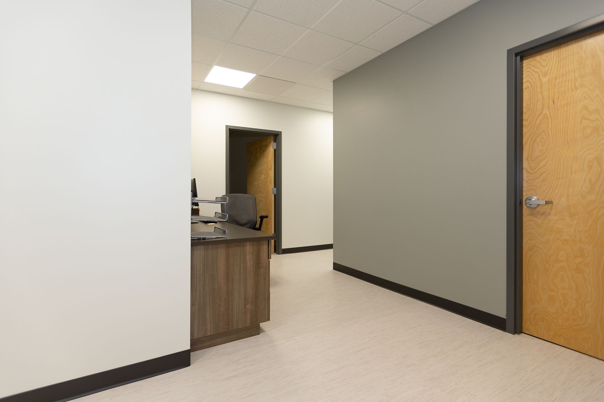 Empty office hallway with white walls, beige floor, wooden door, and reception desk opening on the left