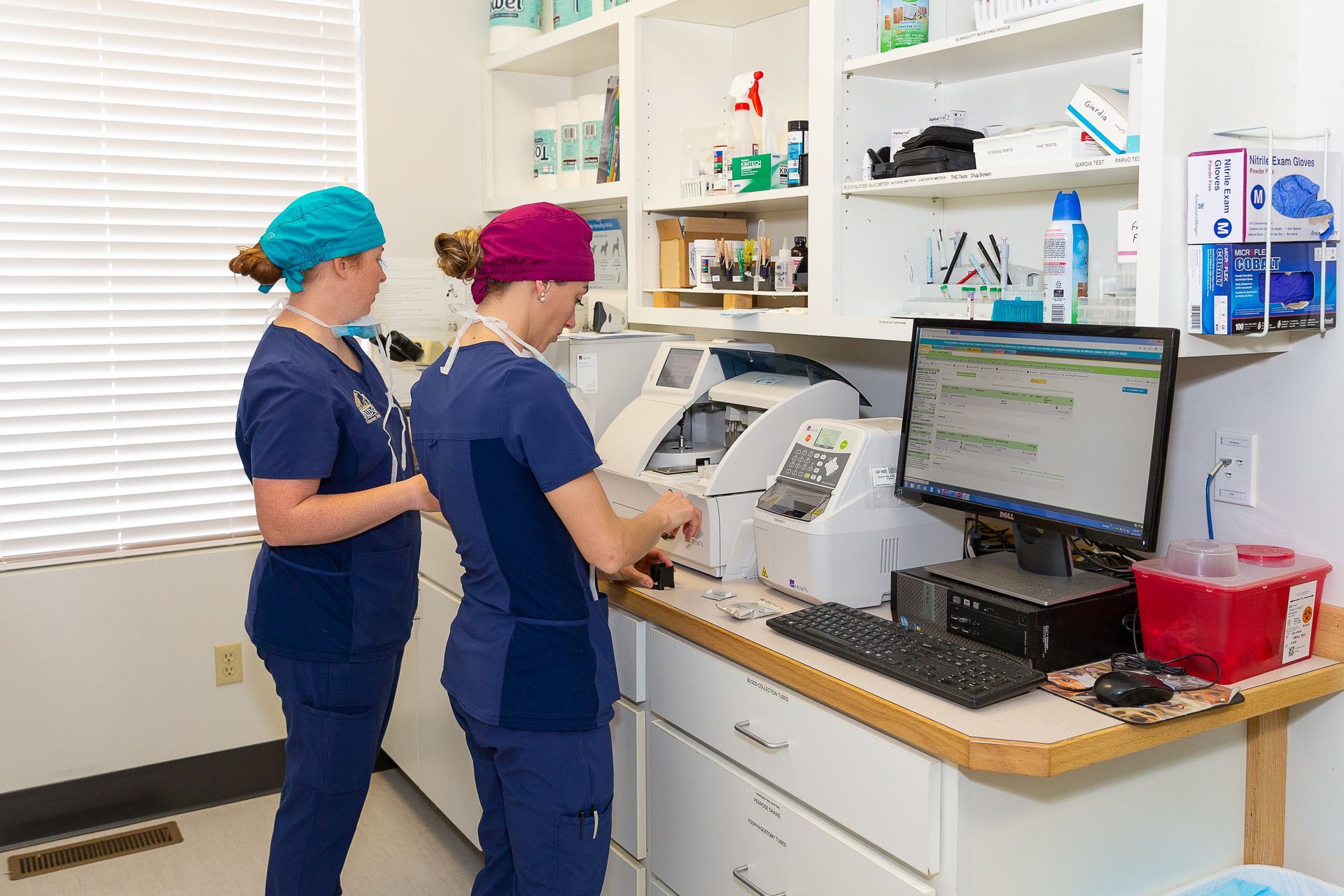 Two lab workers in blue scrubs operate medical equipment at a clinic counter with a computer screen.