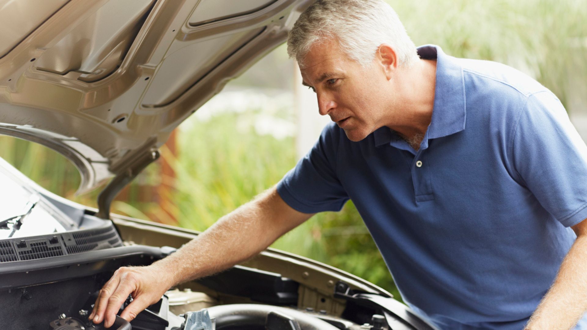 Man in a blue polo checks a car engine with the hood open outdoors