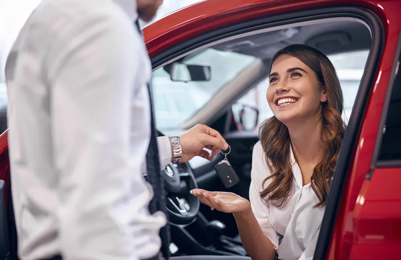 Woman smiling and handing car keys to a person beside a red car