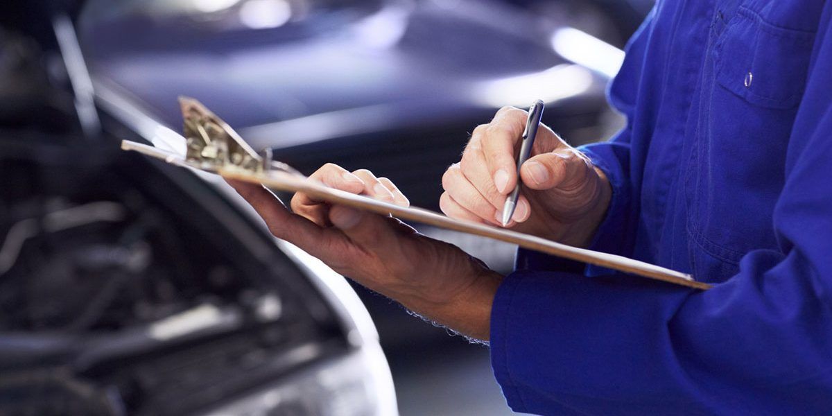 Mechanic in blue uniform writing on a clipboard beside a car engine