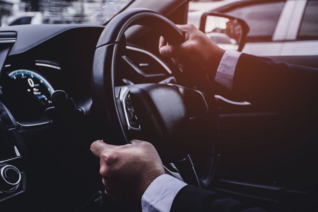 Mani che stringono il volante di un'auto, con il cruscotto visibile in penombra.