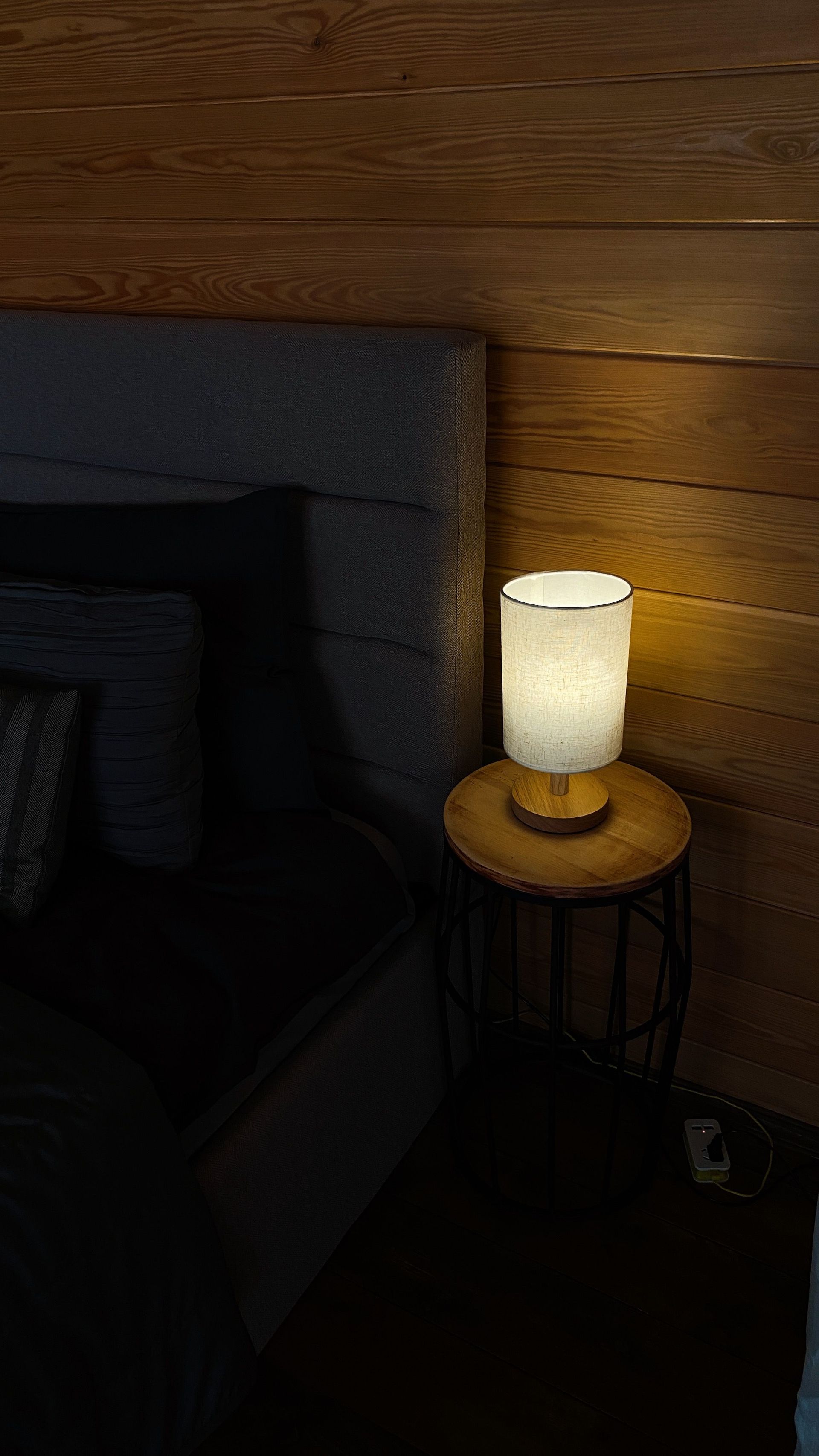 Warm bedside lamp on a wooden side table beside a gray bed in a dimly lit bedroom