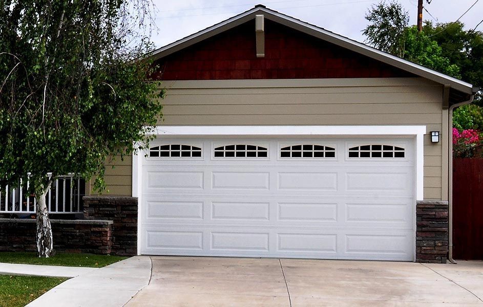 A single-story garage with a white sectional door featuring arched windows, beige siding, and stone-accented corners.