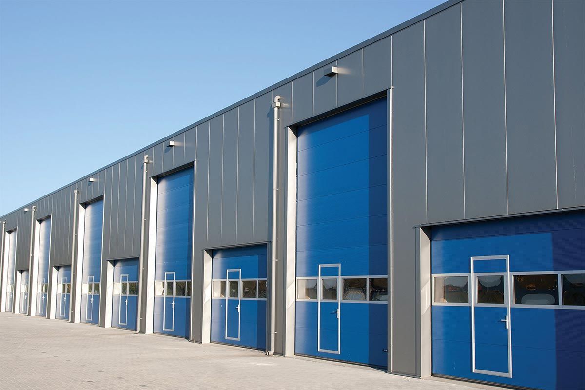 Exterior of a large industrial warehouse with multiple blue industrial roll-up doors against a grey metal wall.