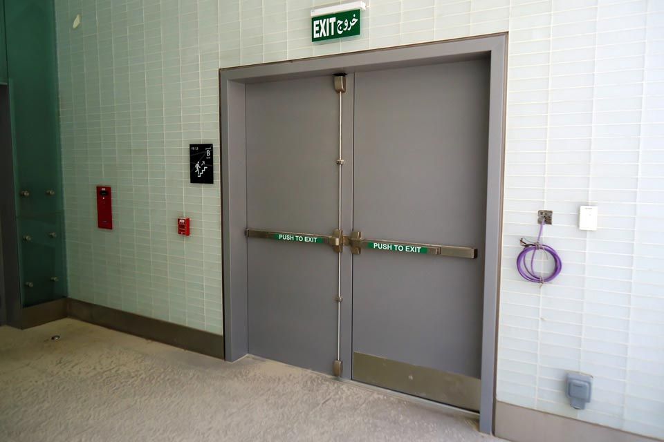 Double grey emergency exit doors with crash bars under an illuminated exit sign in a tiled corridor.