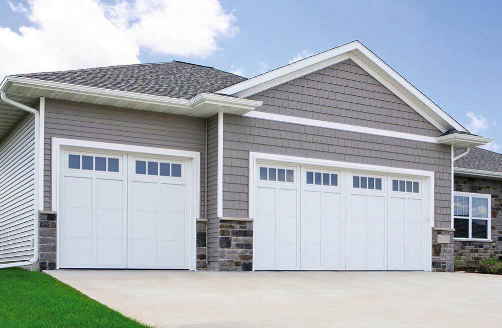 A suburban house with a three-car garage, white doors with windows, grey siding, stone accents, and a grey shingled roof.