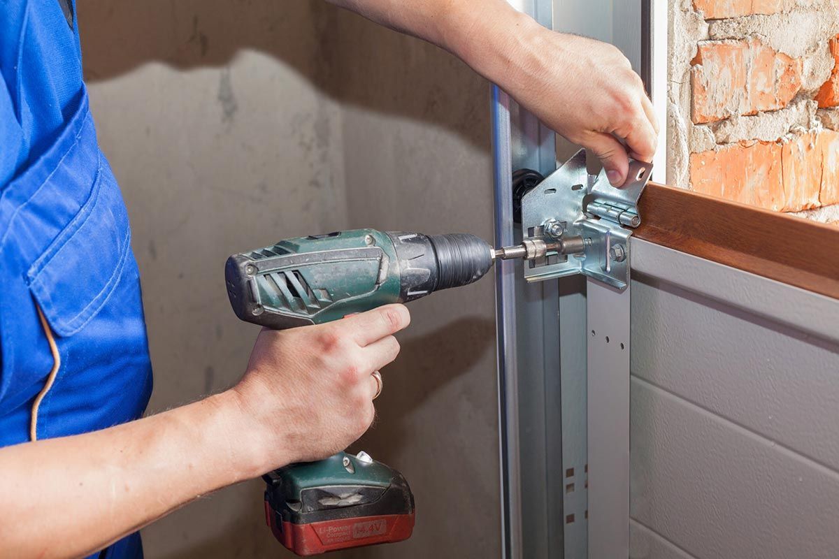A person in blue workwear uses a power drill to install a metal hinge onto a garage door panel next to a brick wall.
