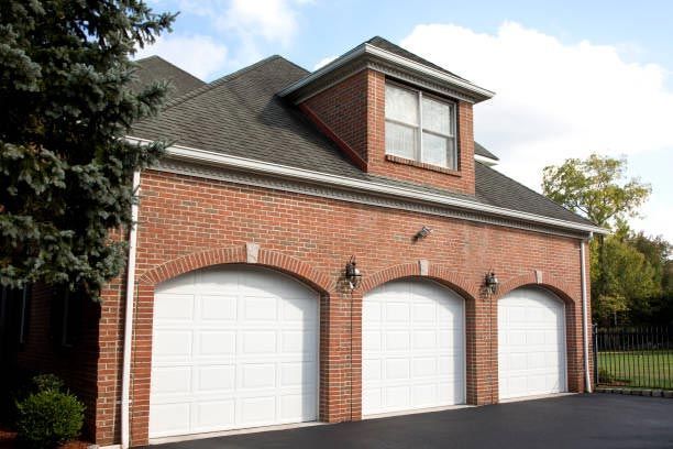 A brick, three-car garage featuring arched white doors and a dormer window under a gray shingled roof.