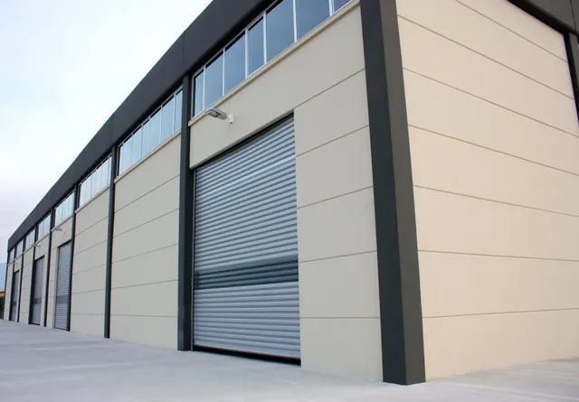 A modern industrial warehouse building with beige walls, dark trim, and multiple large metal roller shutter doors.