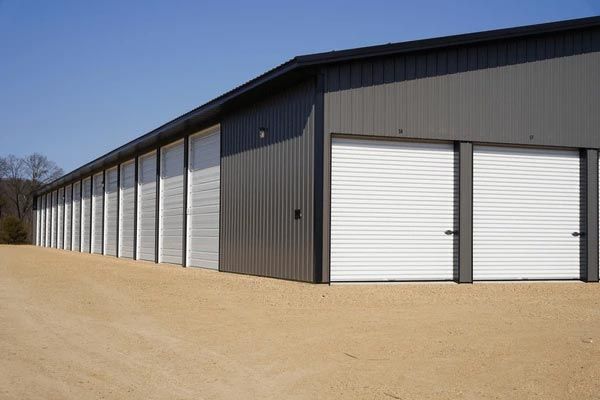 A long, dark gray metal self-storage building with rows of white roll-up doors on a gravel lot under a clear blue sky.