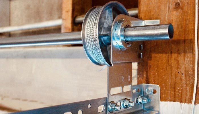 A close-up of a garage door cable drum mounted on a torsion bar, attached to a metal bracket on a wooden frame.