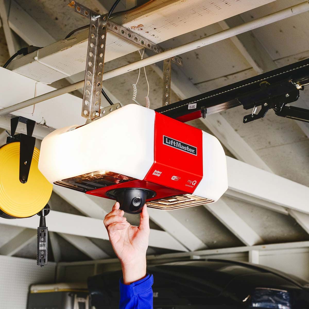 A hand adjusting the camera on a red and white LiftMaster garage door opener mounted on a wooden garage ceiling.