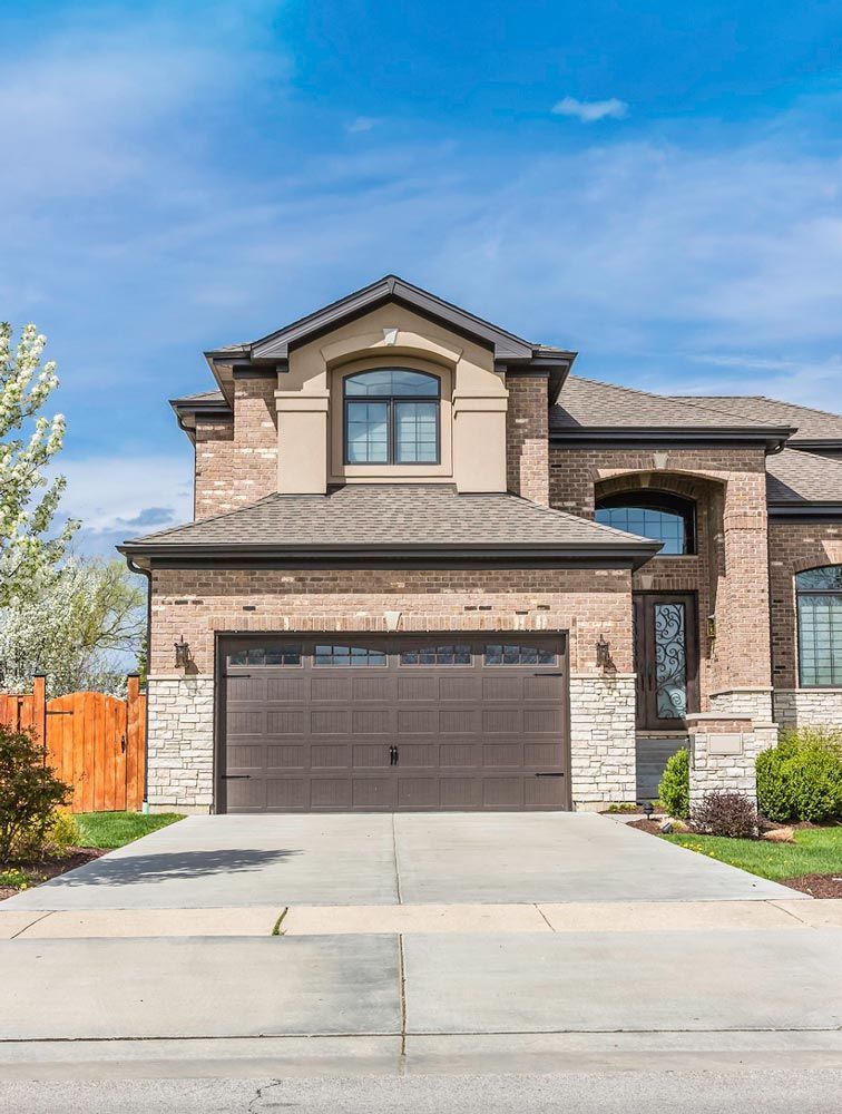 A two-story tan brick suburban house with a brown garage door, arched entryway, and concrete driveway under a blue sky.