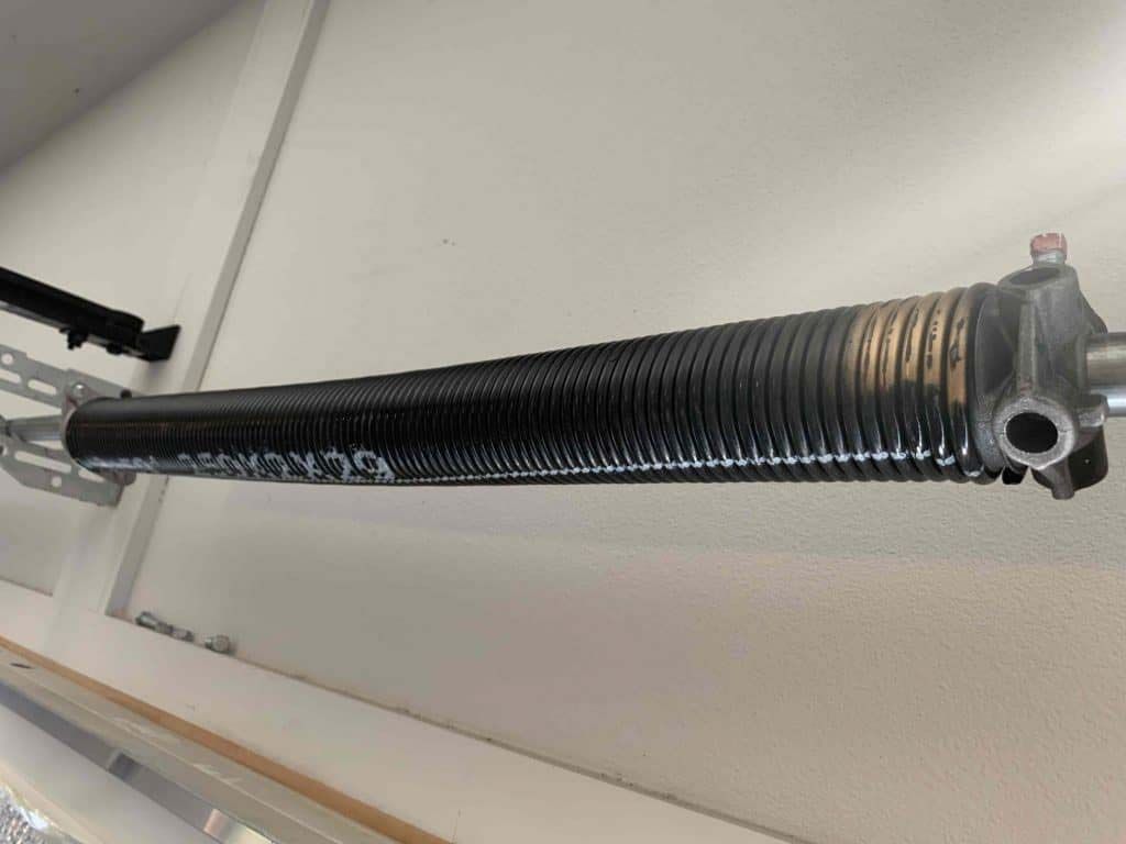 A black metal garage door torsion spring mounted horizontally on a support bracket against a white wall.