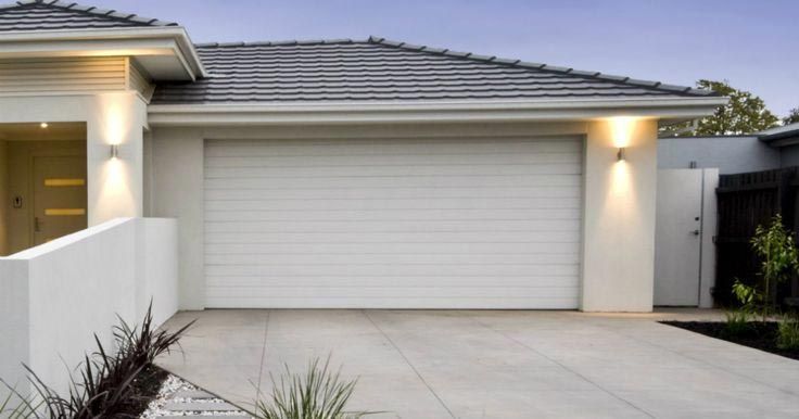 A modern house with a large white garage door, smooth concrete driveway, and exterior wall-mounted lighting.