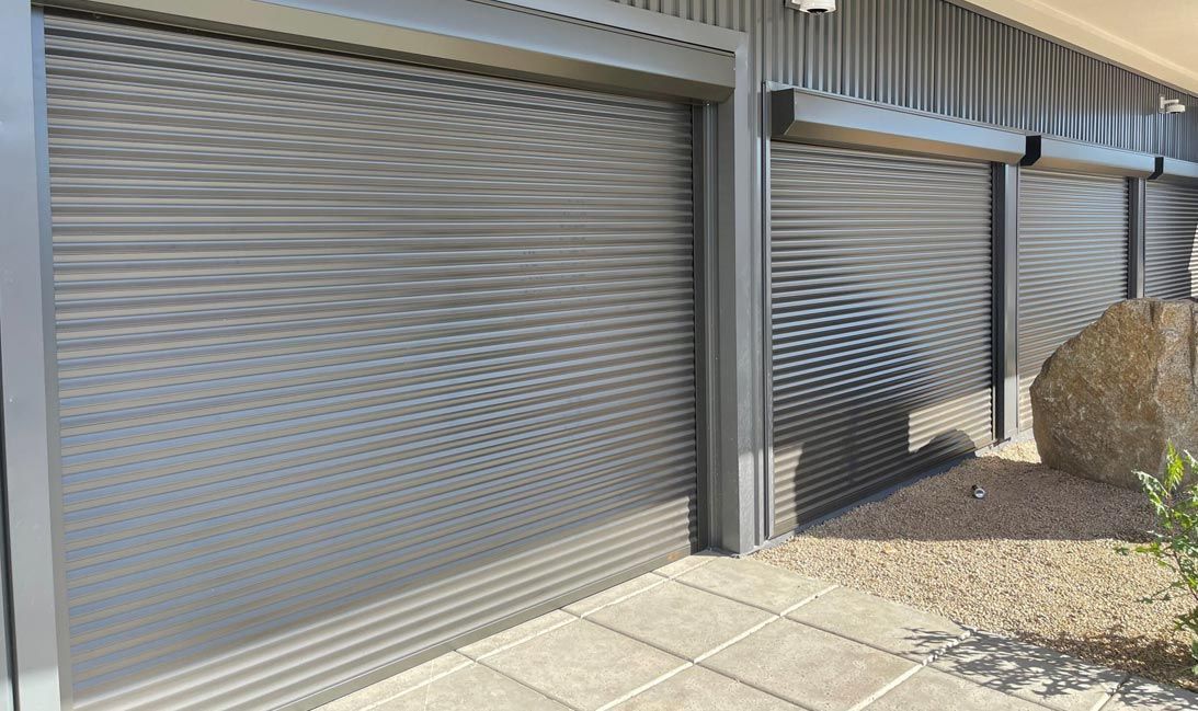 Modern dark grey metal roller shutters covering a garage door and adjacent windows on a building exterior.
