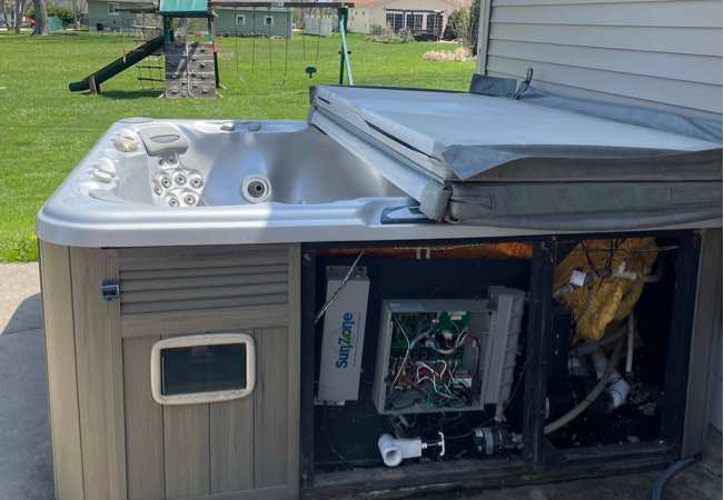 a gray hot tub missing a panel with an open cover in front of a playset