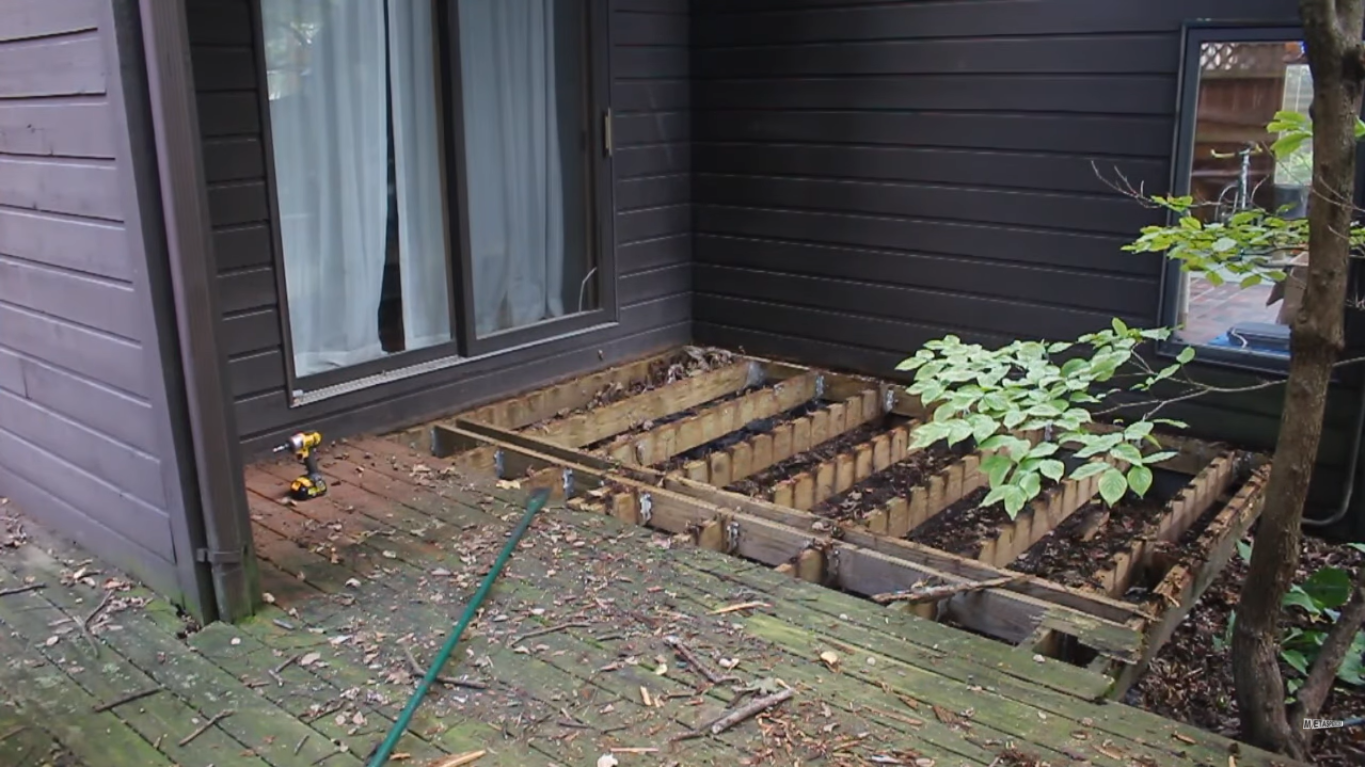 recently removed wooden deck with exposed beams. Corroded moldy green stained deck and the side of a house are also visible