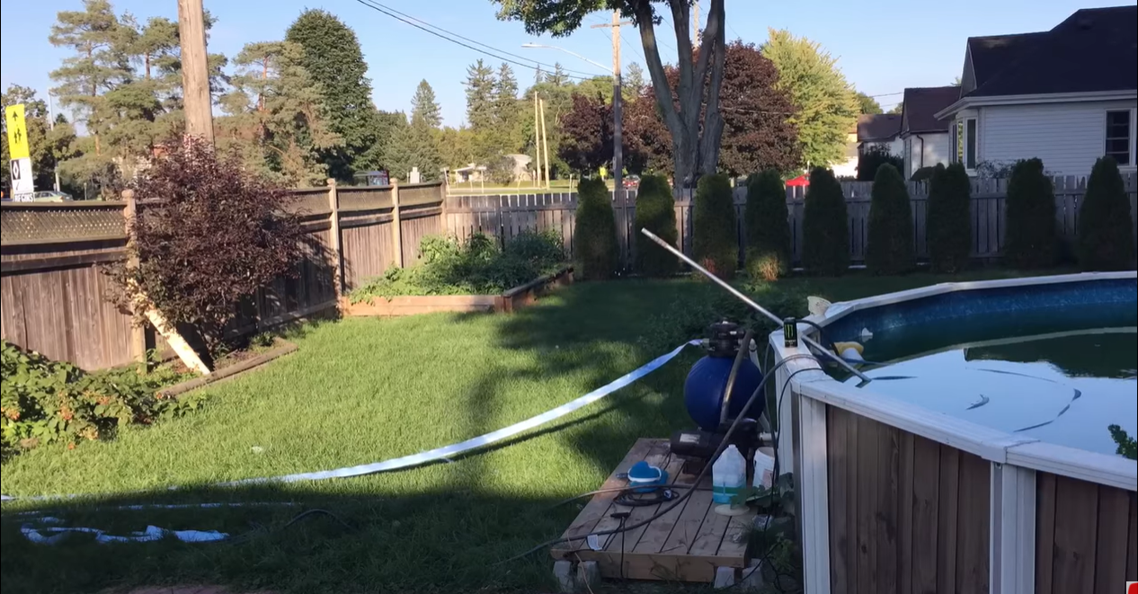 View of a backyard. Above ground pool visible on the right hand side. Small trees visible in the background lined up next to a fence. Small boxed garden in the back left corner of the photo