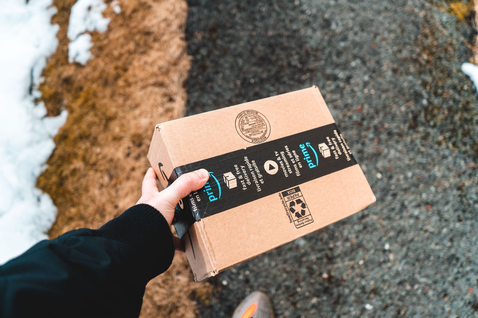 Hand holding a cardboard package with a black shipping label on a snowy outdoor path