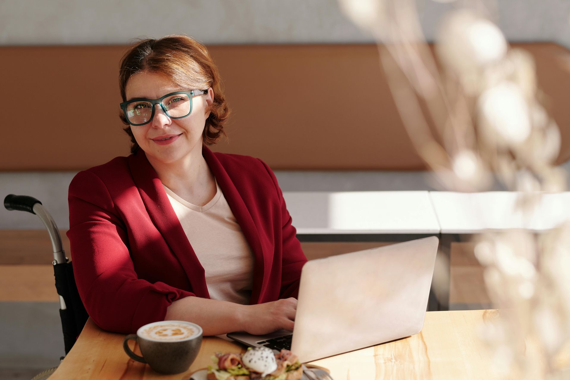 Woman in a red blazer smiling at a laptop in a sunlit café, with coffee on the table.