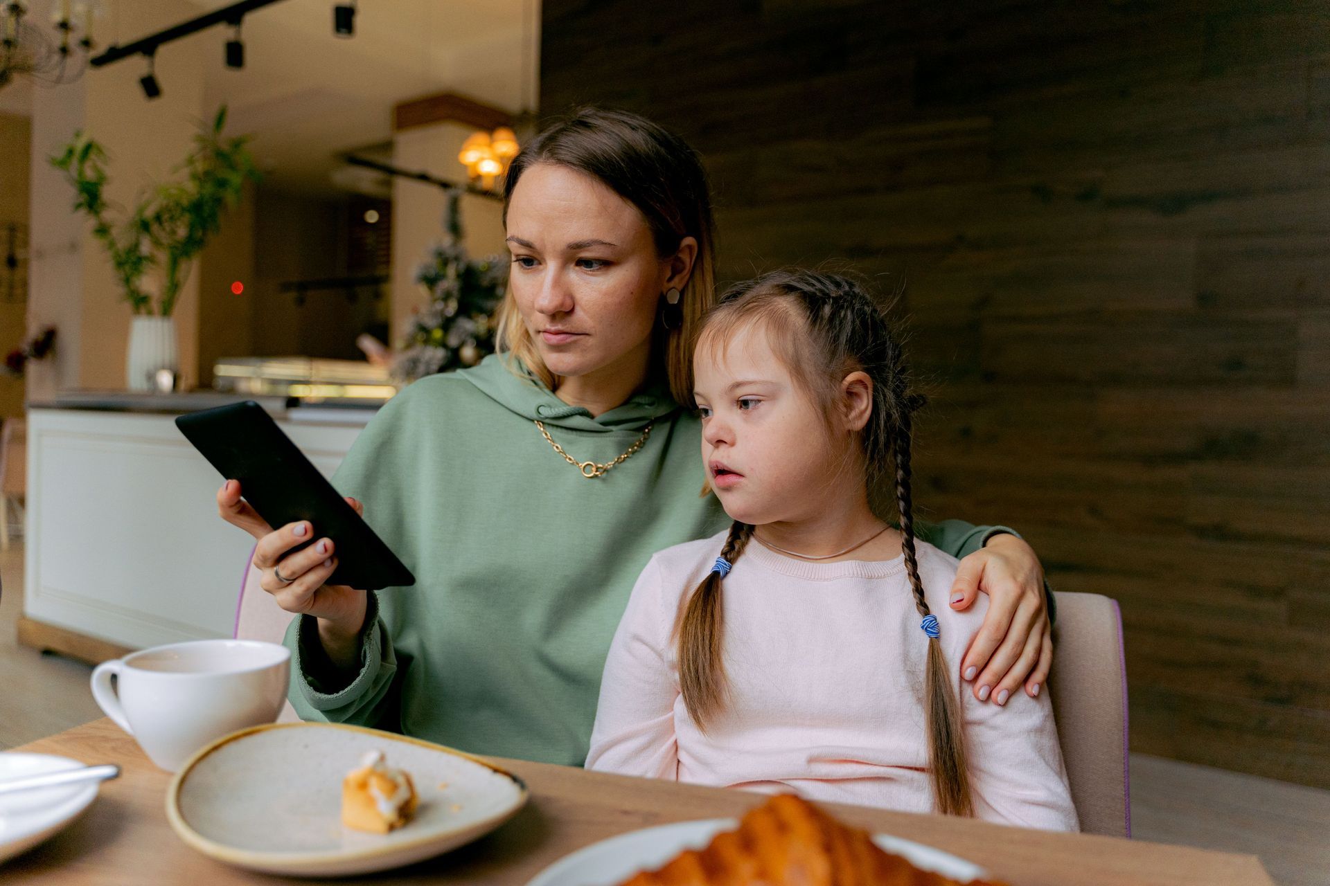 Adult and child at a cafe table, looking at a tablet beside cups and pastries