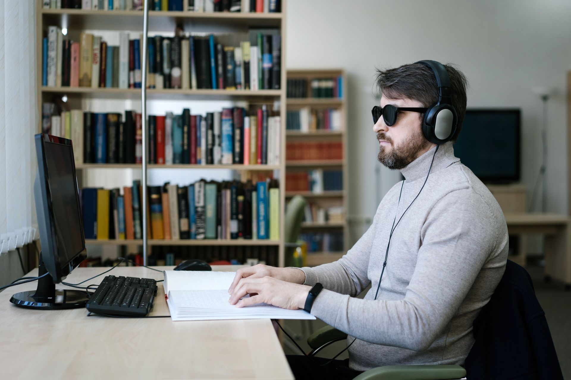 A man wearing headphones and sunglasses types at a desk in a home office with bookshelves.